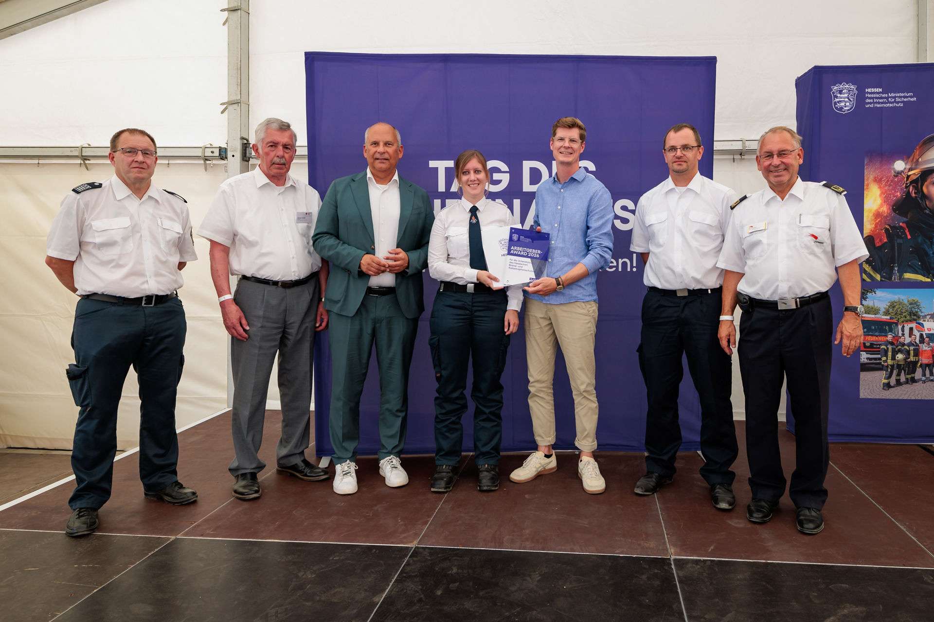 Sieben Personen stehen bei einer Preisverleihung in einem Veranstaltungszelt vor einem violetten Hintergrund. Zwei Personen in der Mitte halten gemeinsam eine Urkunde mit der Aufschrift „Arbeitgeber-Award 2026“, mehrere Personen tragen Uniform.