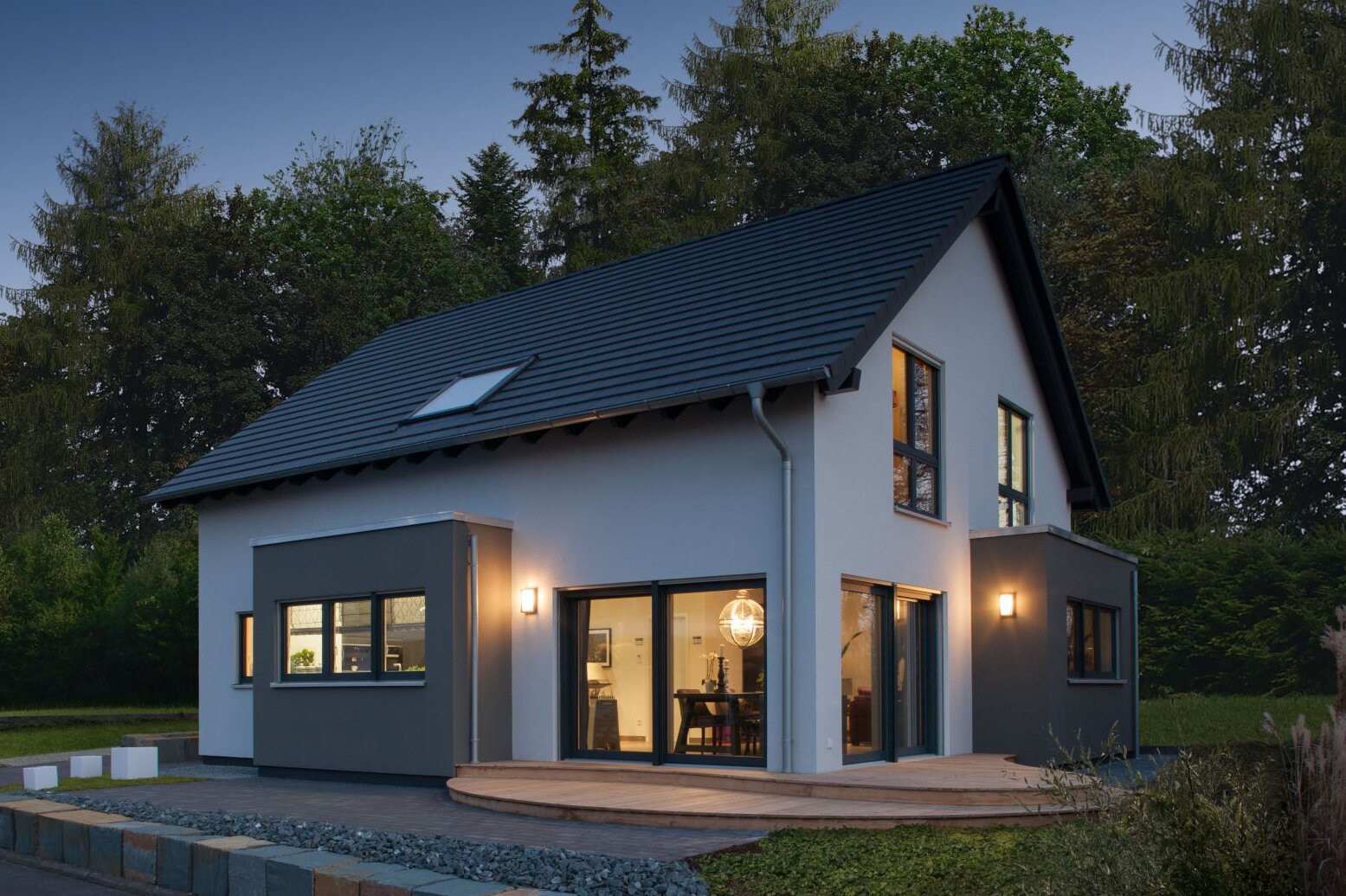 Modernes Musterhaus bei Dämmerung mit anthrazitfarbenem Satteldach, hellgrauer Fassade und großen Fensterflächen. Innen ist warmes Licht sichtbar, außen eine kleine Holzterrasse mit Kiesfläche und Natursteinmauer.