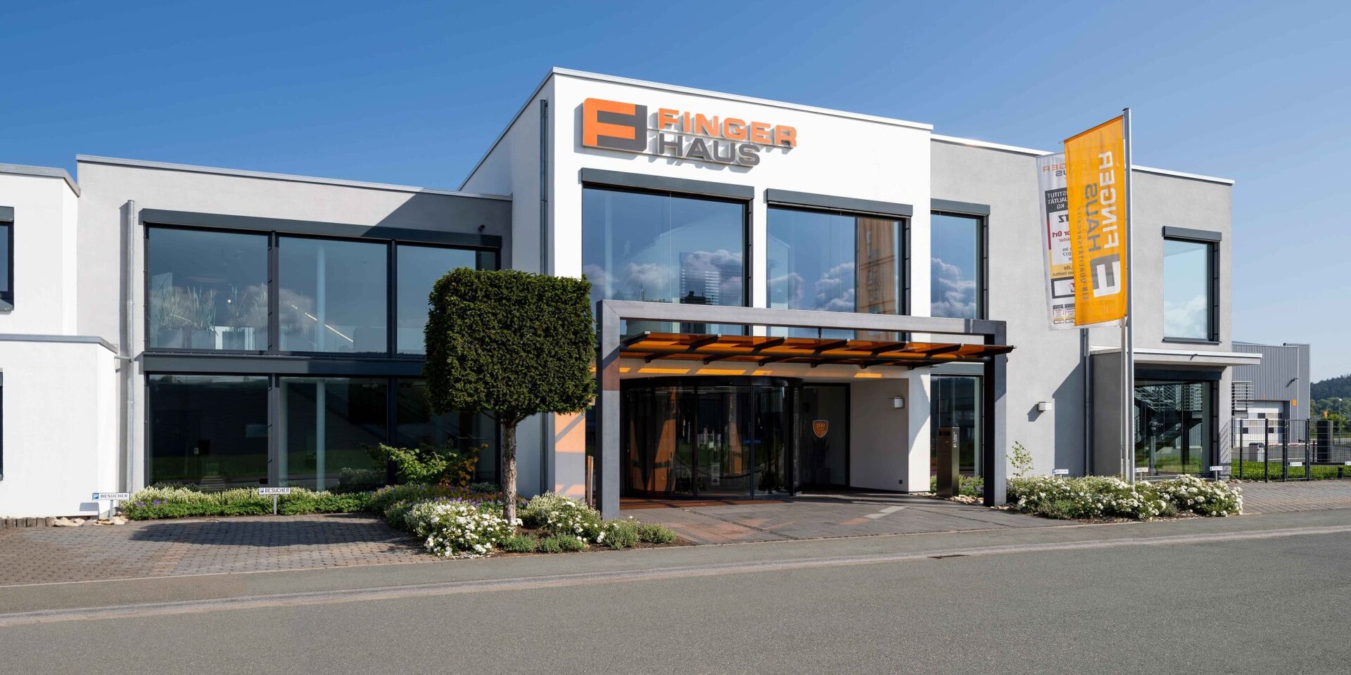 FingerHaus Verwaltungsgebäude mit großen Fenstern, Flachdach und orange-weißem FingerHaus-Logo über dem Eingang, davor ein rechteckig geschnittener Baum und eine orange-weiße Fahne mit Schriftzug