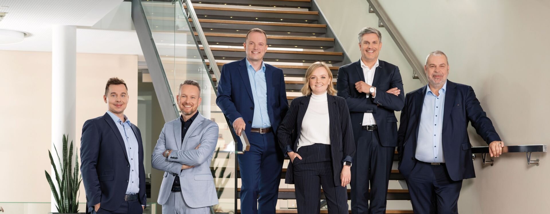 Gruppe von Kollegen aus dem Vertrieb von FingerHaus im Verwaltungsgebäude am Firmenstandort Frankenberg, stehend auf einer Treppe im Innenraum, Businesskleidung, modernes Treppenhaus mit Glasgeländer und Holzstufen.