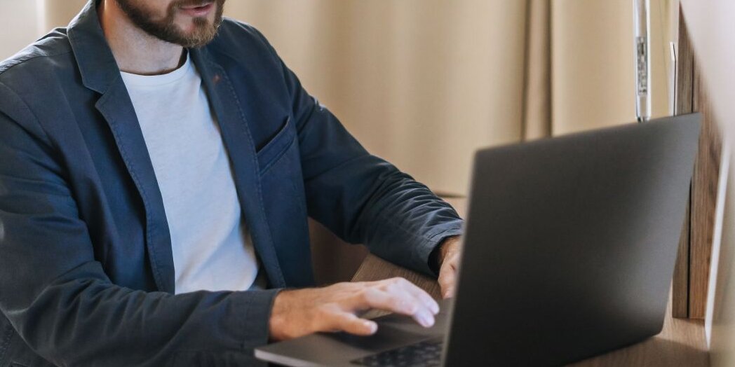 Sitzender Mann an einem Schreibtisch im Innenraum, arbeitet an einem Laptop, Tischlampe mit Gelenkarm, Smartphone auf Holzschreibtisch, Vorhang und Fenster im Hintergrund.