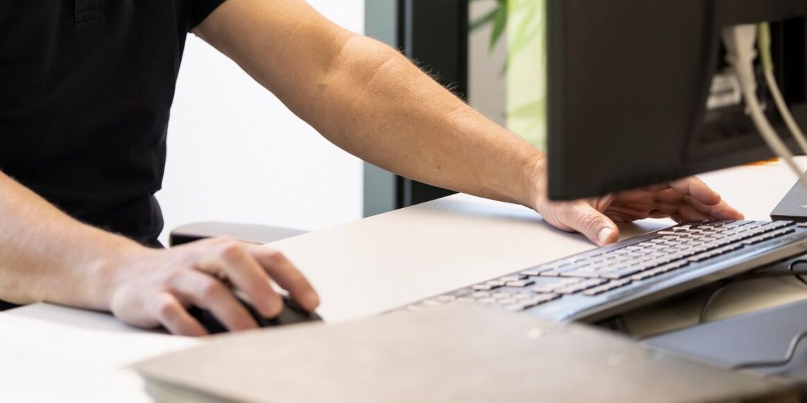 Ein Mann sitzt an einem Schreibtisch vor einem Computerbildschirm und trägt ein Headset. Er arbeitet konzentriert und lächelt, während er mit Maus und Tastatur interagiert.