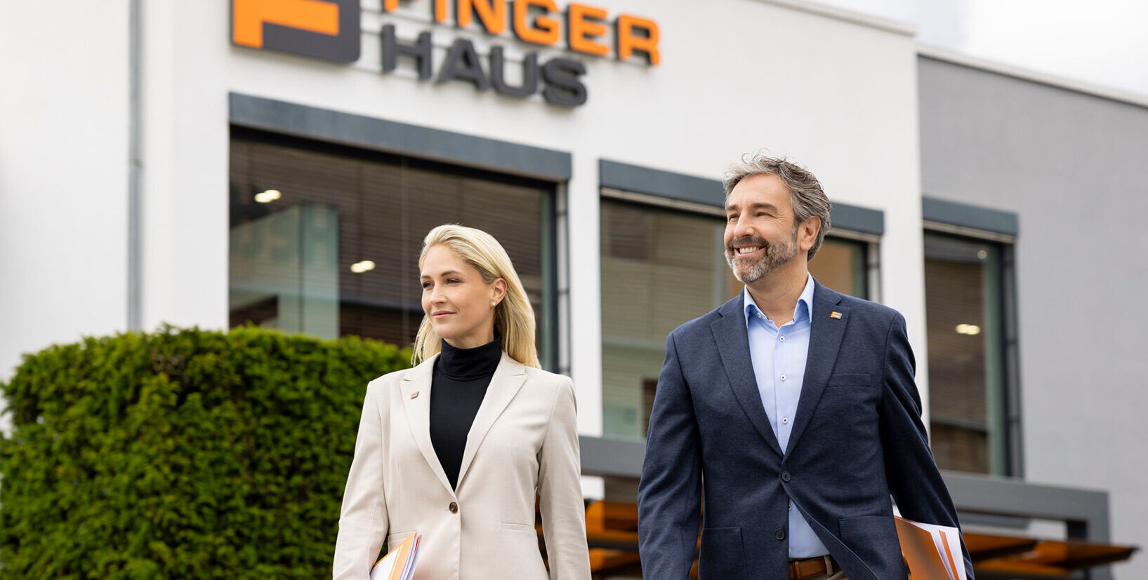 Zwei Fachberater von FingerHaus gehen nebeneinander vor dem Firmengebäude, Businesskleidung, jeweils mit Unterlagen in der Hand, FingerHaus-Logo an der Gebäudefassade im Hintergrund, Außenbereich bei Tageslicht.