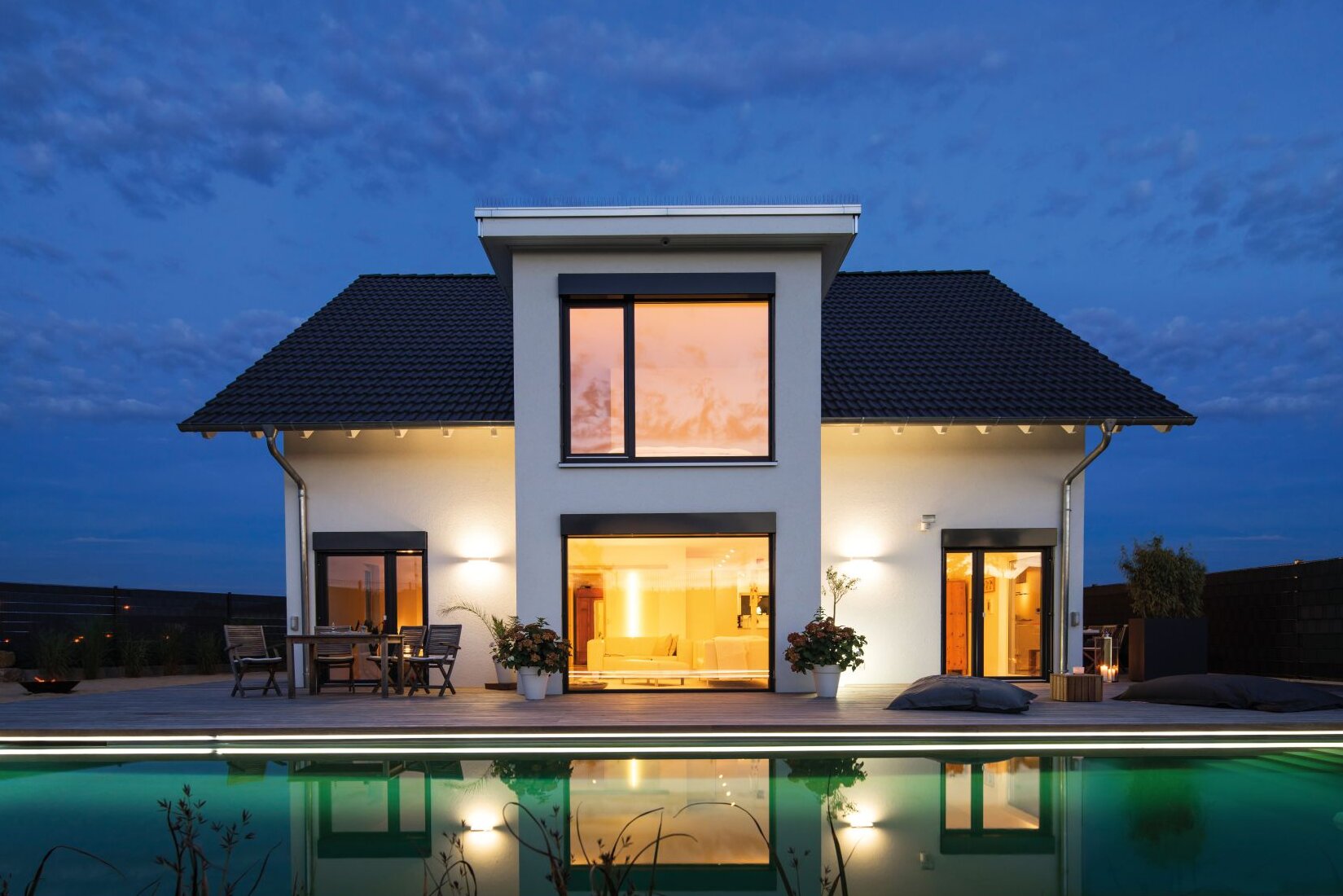 Zweigeschossiges Einfamilienhaus des Hauskonzepts VIO von FingerHaus mit Satteldach, weißer Putzfassade, zentralem Fassadenvorsprung, großen bodentiefen Fenstern, beleuchtetem Innenraum und Pool im Vordergrund bei Dämmerung.