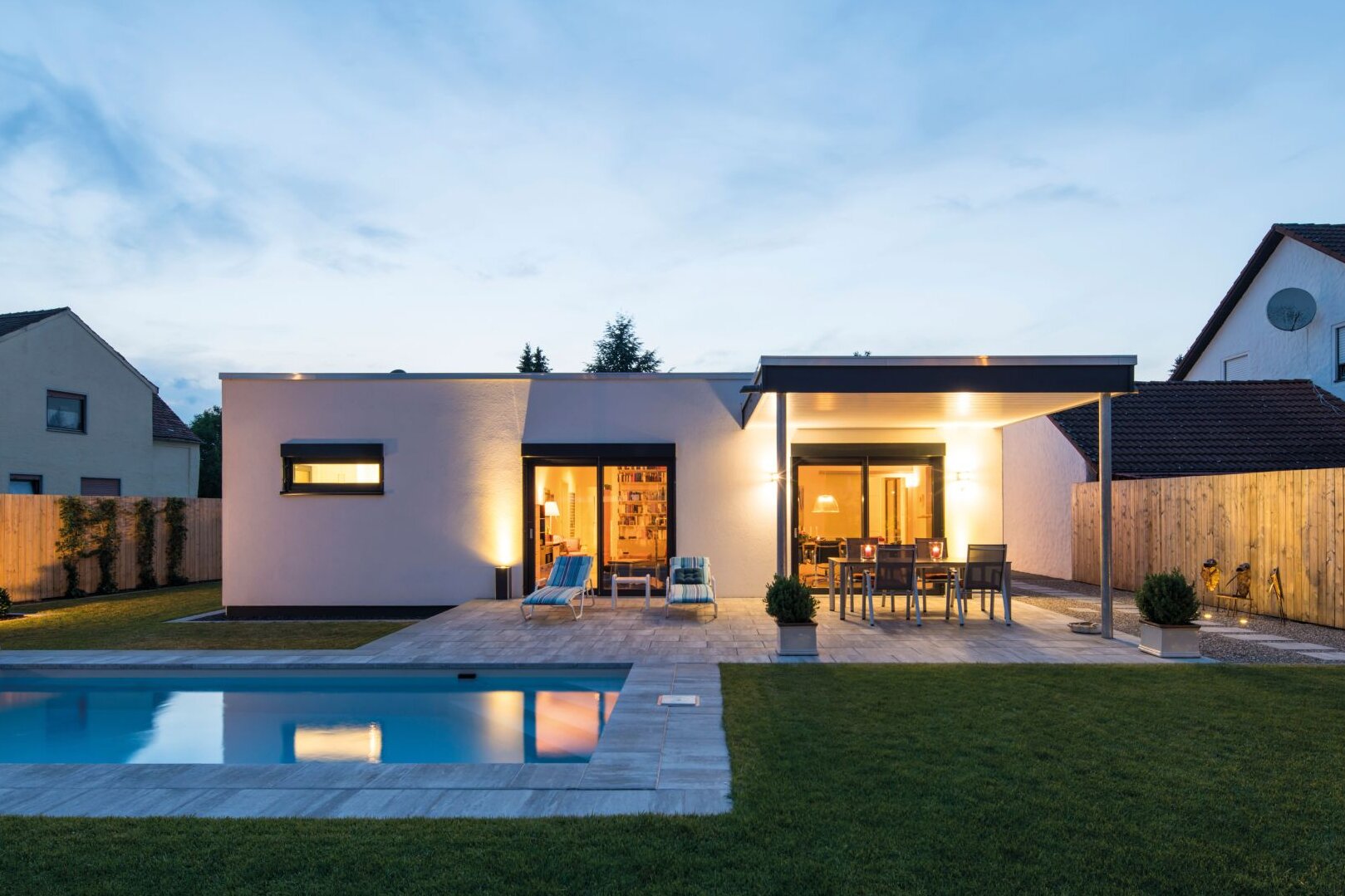 Moderne, eingeschossige Architektur des Hauskonzepts NIVO von FingerHaus mit Flachdach, weißer Putzfassade, großer Terrasse mit Überdachung, bodentiefen Fenstern und beleuchtetem Innenraum, Pool und Rasenfläche im Vordergrund bei Dämmerung.