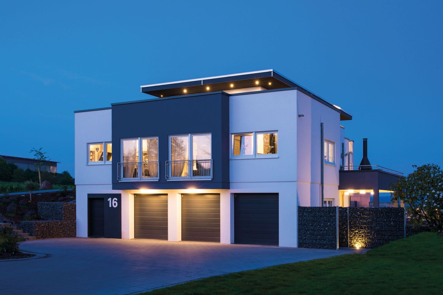 Modernes, frei geplantes Finger-Haus mit Flachdach bei Dämmerung, zweigeschossige kubische Architektur, weiße und dunkel abgesetzte Fassadenflächen, Balkone mit Glasgeländer, drei Garagentore, beleuchteter Innenraum und gepflasterte Einfahrt im Vordergrund.