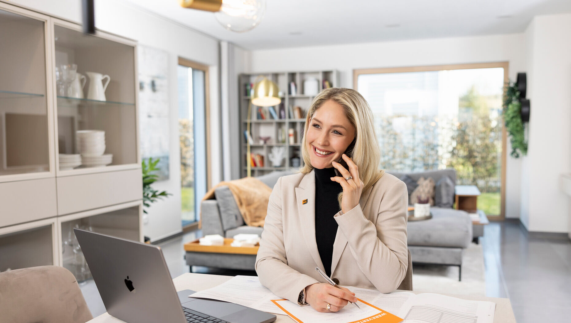 Vertriebskollegin von FingerHaus sitzt an einem Tisch im Wohnbereich, telefoniert mit einem Kunden, Laptop geöffnet, Unterlagen und Schreibblock vor sich, Stift in der Hand, moderne Wohnraumausstattung im Hintergrund.