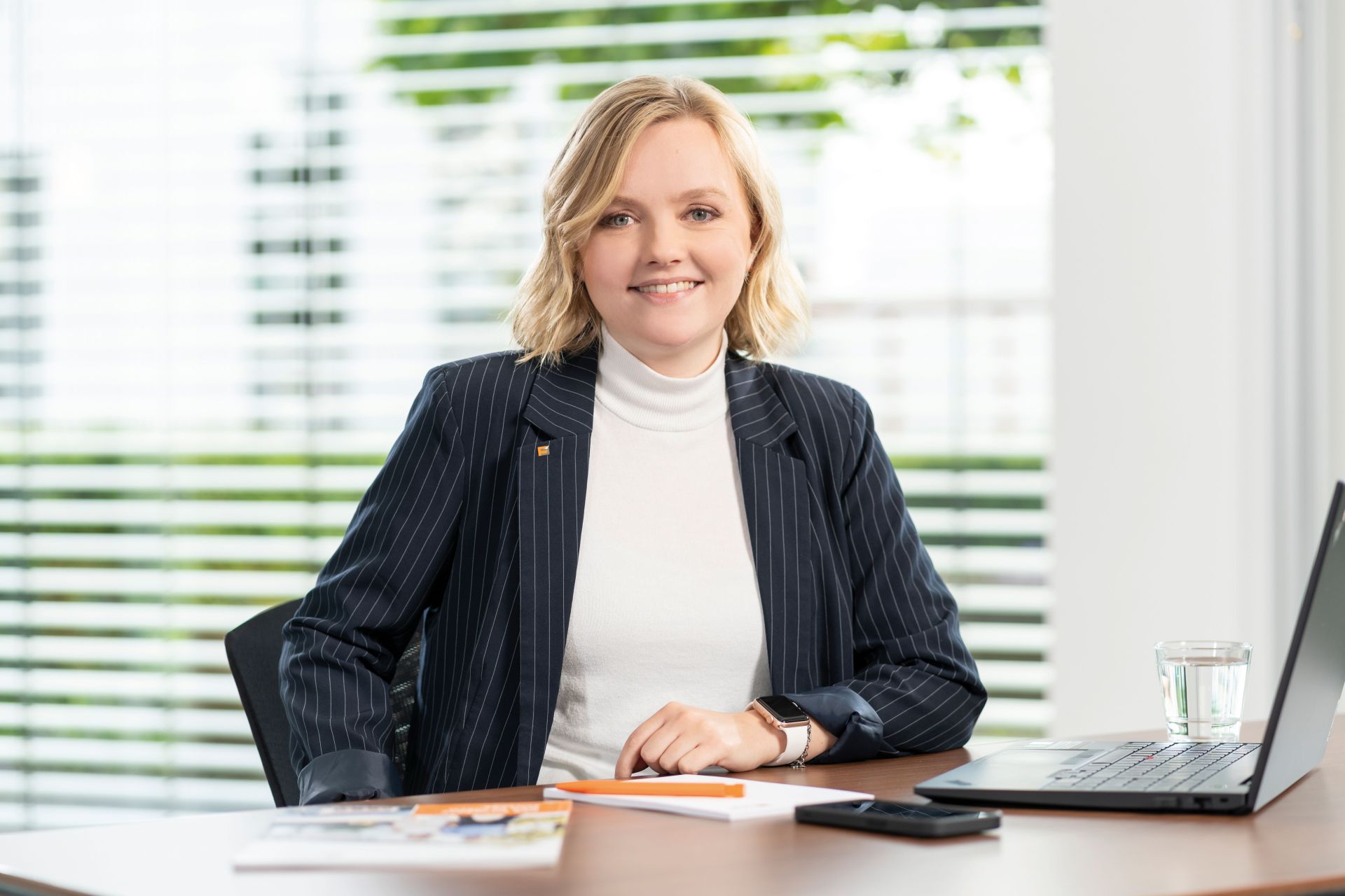 Ansprechpartnerin für Personalrecruiting Vertrieb von FingerHaus sitzt an einem Schreibtisch im Büro, Blick in die Kamera, Businessanzug mit Nadelstreifen und hellem Rollkragenpullover, Laptop, Smartphone, Notizblock und Glas Wasser auf dem Tisch, Jalousien im Hintergrund.
