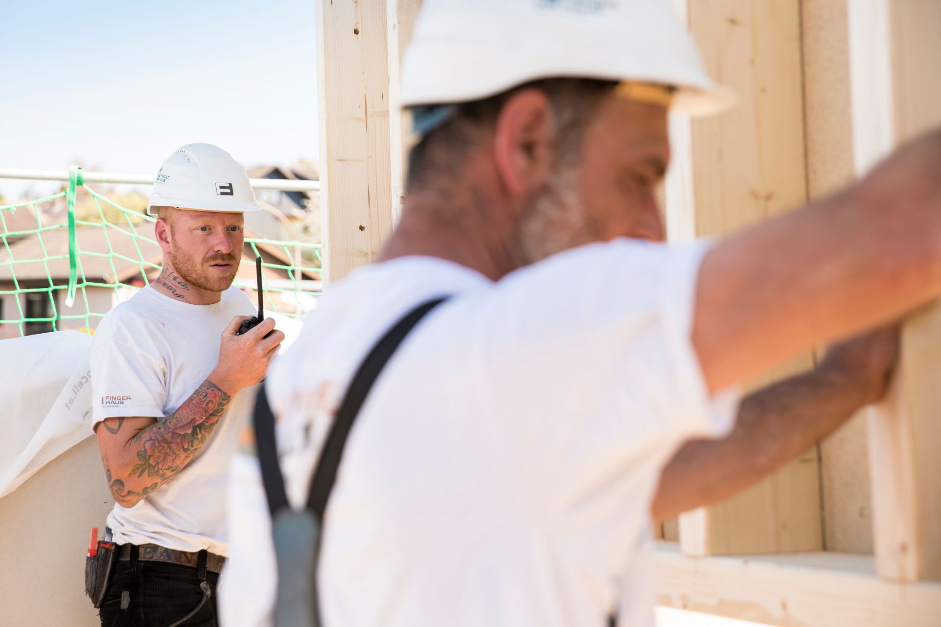 Zwei Bauarbeiter auf einer Baustelle im Holzrohbau, beide mit Schutzhelm, eine Person im Vordergrund bei der Montage eines Holzbauteils, eine weitere Person im Hintergrund mit Funkgerät, Holzständerwerk und Sicherungsnetz sichtbar.