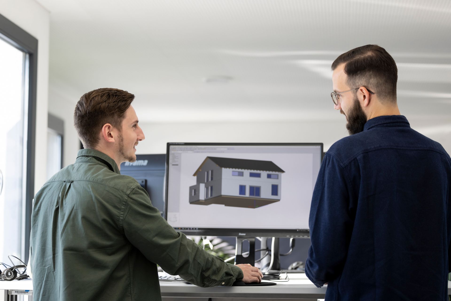 Zwei Bauzeichner von FingerHaus im fachlichen Austausch vor einem Monitor mit einem digitalen Hausmodell.