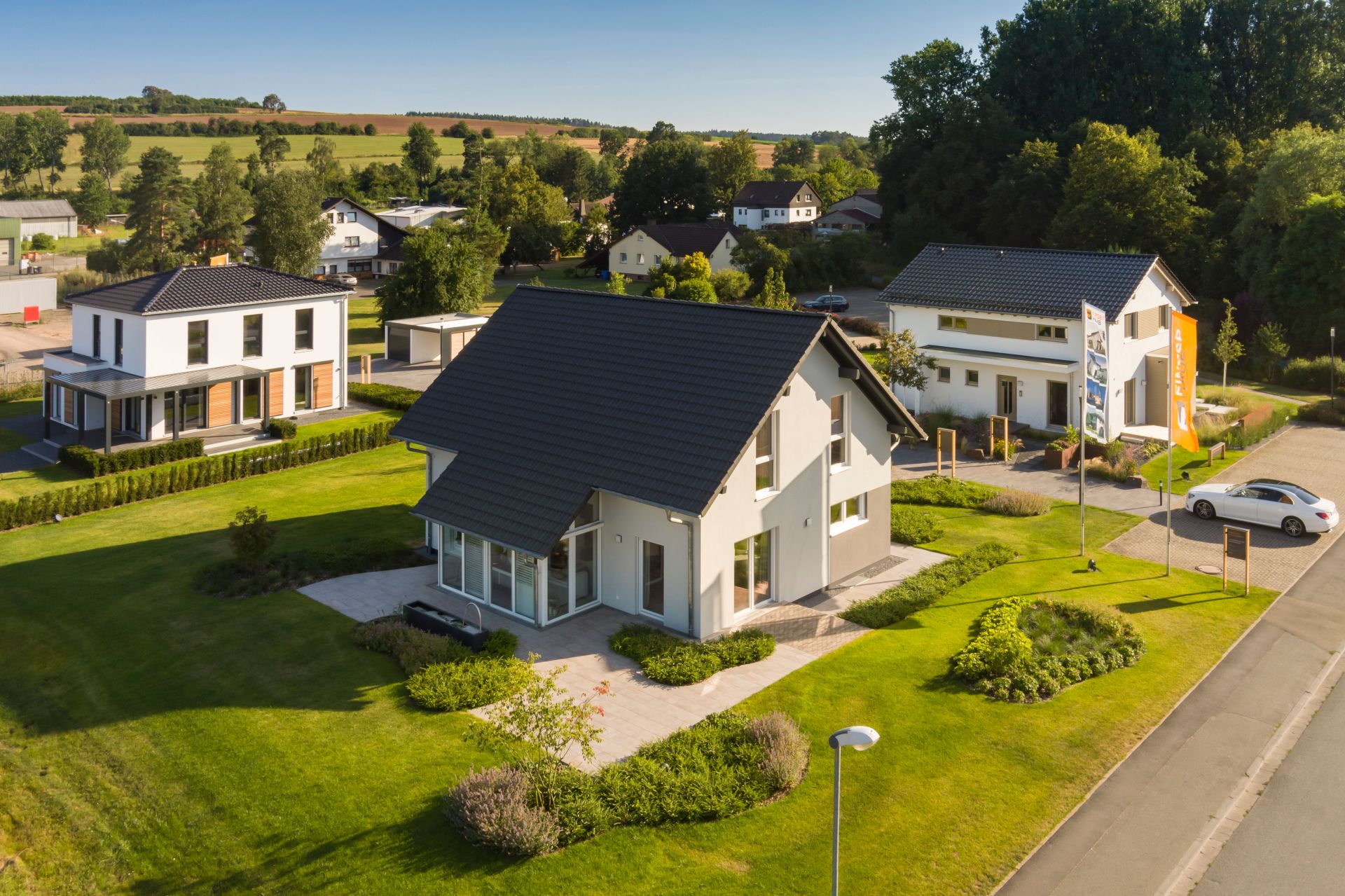 Luftaufnahme des Musterhausparks von FingerHaus in Frankenberg mit mehreren modernen Musterhäusern, gepflegten Grünflächen und Flaggen. Im Hintergrund sind Felder, Bäume und Wohnhäuser der Umgebung zu sehen.