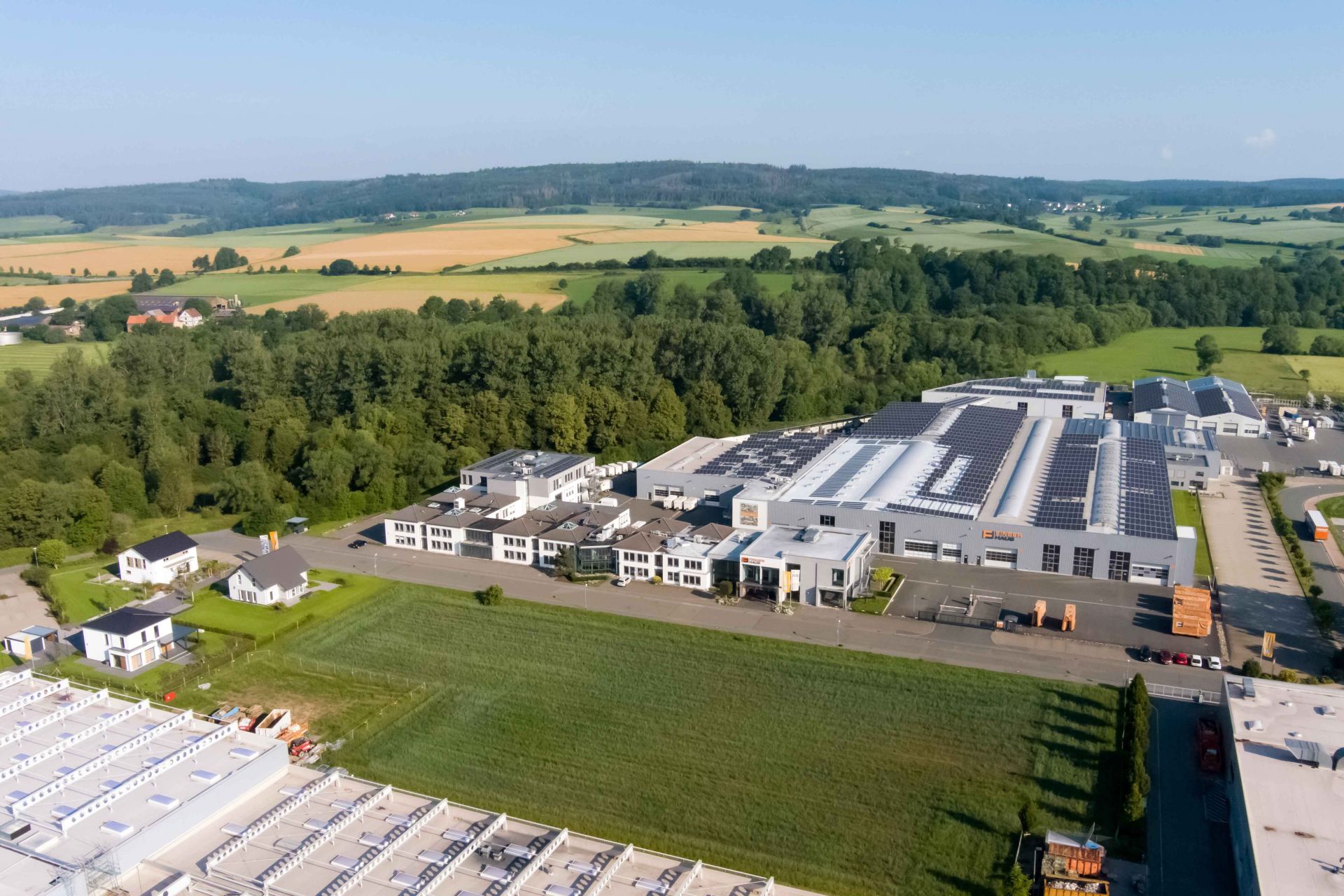 Luftaufnahme des FingerHaus-Standorts in Frankenberg (Eder) mit großflächigen Produktions- und Verwaltungsgebäuden, hellen Hallendächern mit Photovoltaikanlagen, angrenzenden Grünflächen, Zufahrtsstraßen und umliegender Landschaft.