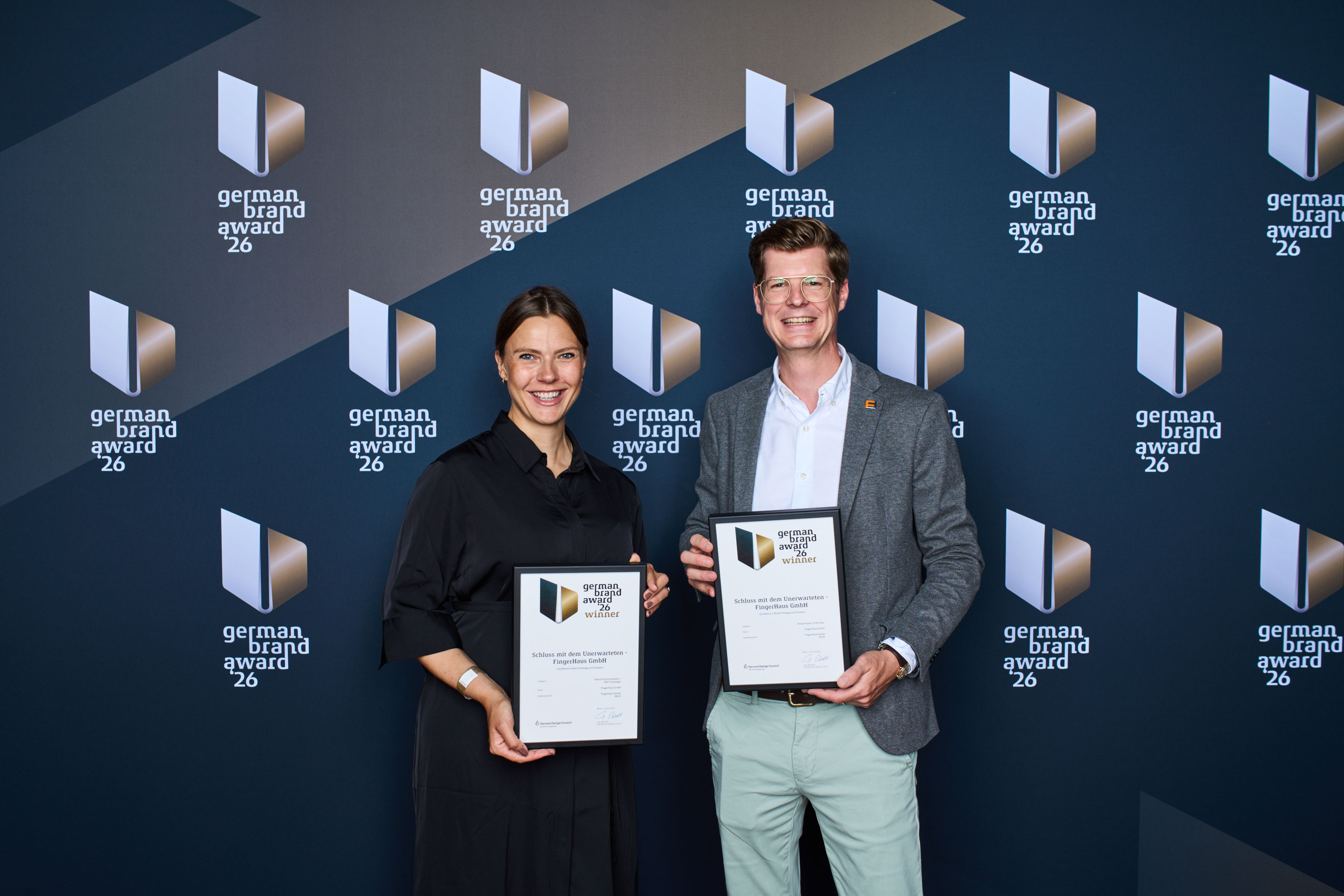 Zwei Personen halten Urkunden des German Brand Award in den Händen und stehen vor einer Veranstaltungswand mit dem Logo des German Brand Award.