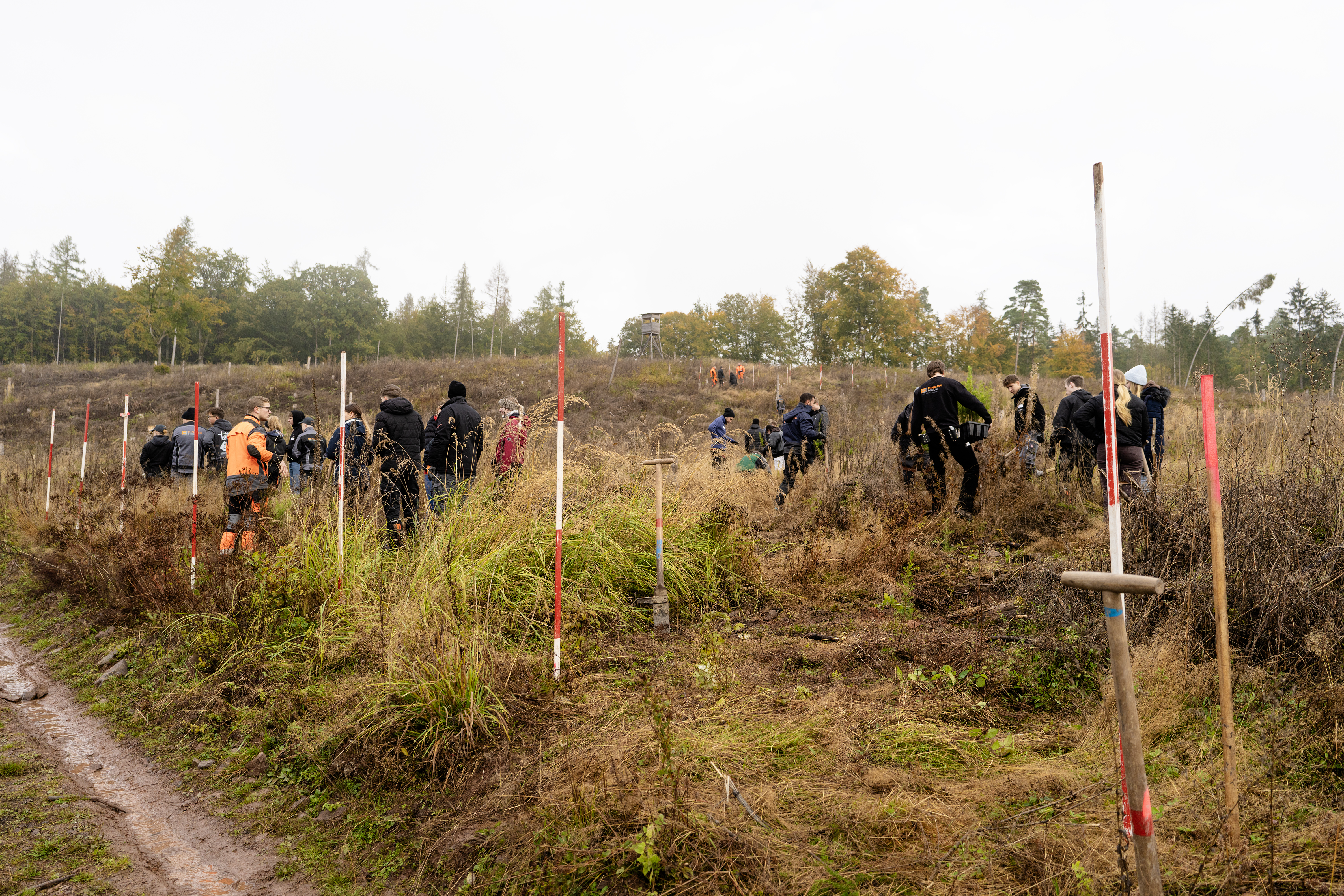 Gruppe von Menschen mit wetterfester Kleidung auf einem unebenen, bewachsenen Gelände mit rot-weißen Markierungsstäben